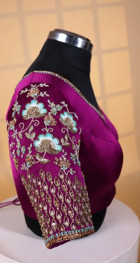 Magenta silk bridal blouse on a dress form, its elbow-length sleeve worked in gold zardosi, sequins and pale blue thread in a lotus-and-leaf pattern above a scalloped beaded cuff