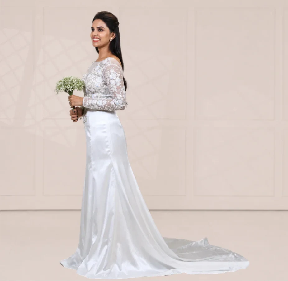 Bride in a custom white bridal gown with a sheer long-sleeved bodice in floral appliqué over an ivory satin trumpet skirt that sweeps into a chapel train, holding a small white bouquet against a pale panelled wall
