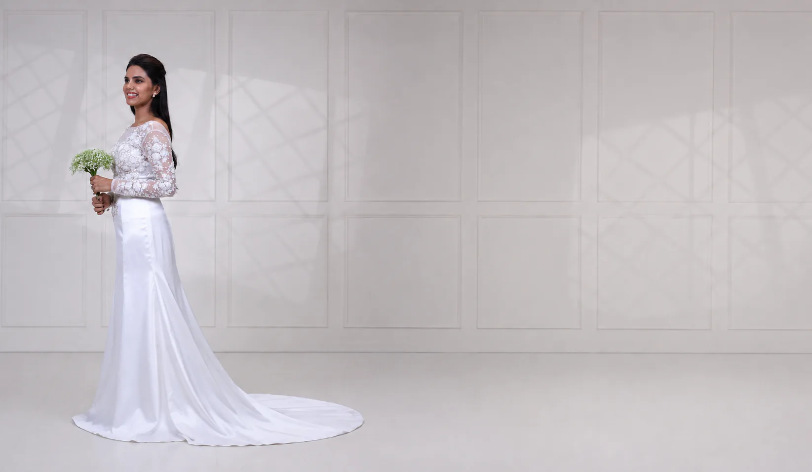 Bride in a custom white bridal gown with a sheer long-sleeved bodice in floral appliqué over an ivory satin trumpet skirt that sweeps into a chapel train, holding a small white bouquet, standing at the left of a wide pale panelled studio wall